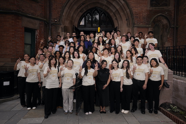 A group of students wearing Vanderbilt University NYC shirts pose together in front of a brick building entrance, making peace signs.